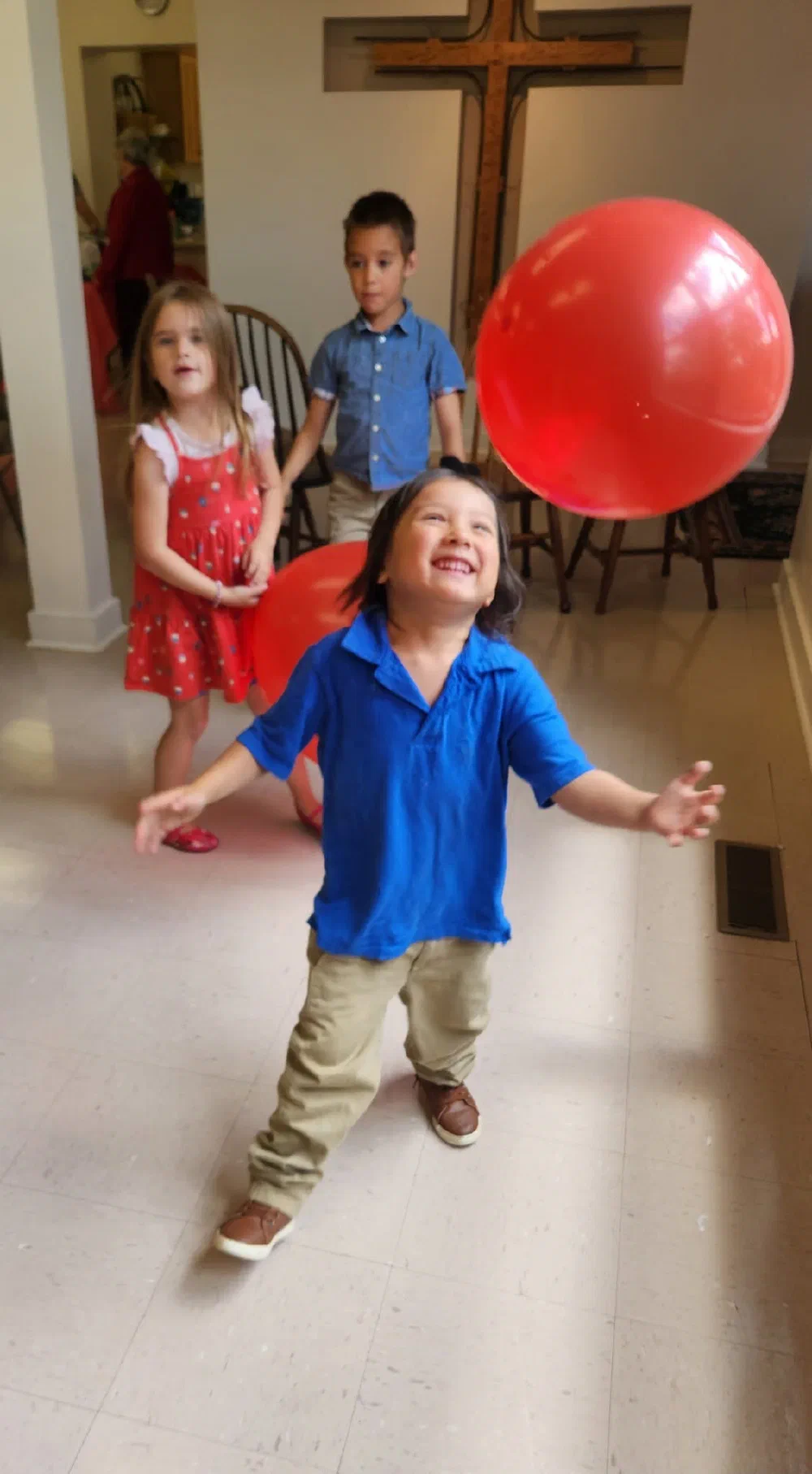 A delighted boy reaches for a red balloon in the parish hall while two friends look on