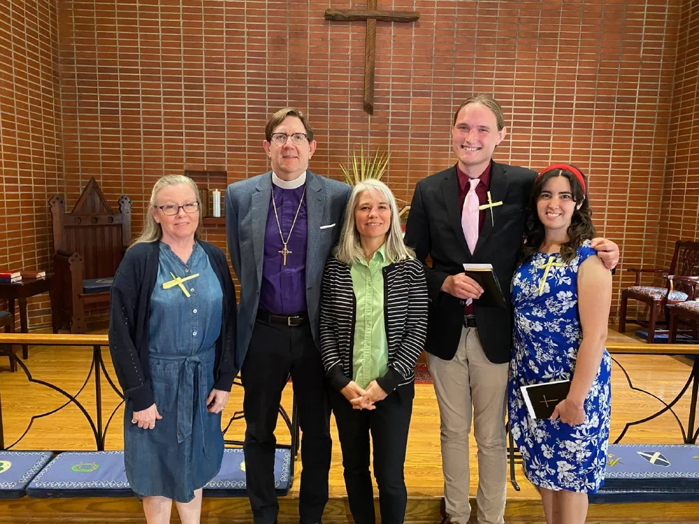 The bishop in a purple clergy shirt poses with newly confirmed adults wearing palm crosses in front of the chancel