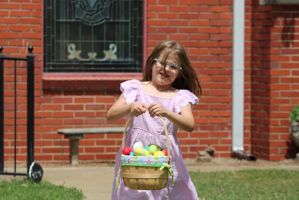 A girl in a pink dress holds a basket full of colorful Easter eggs in front of the church's brick wall