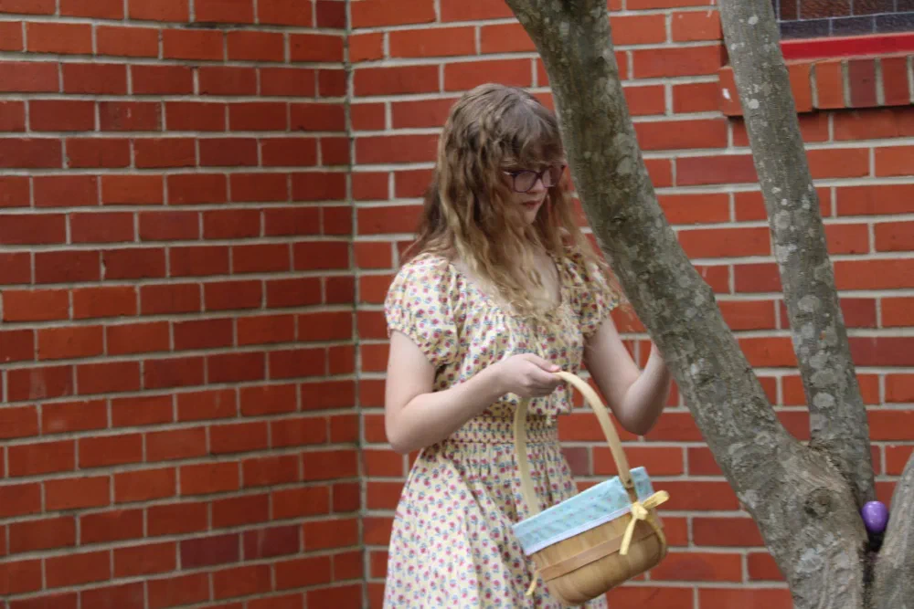 A girl with an Easter basket searches a tree by the brick wall, where a purple egg is tucked in the branches