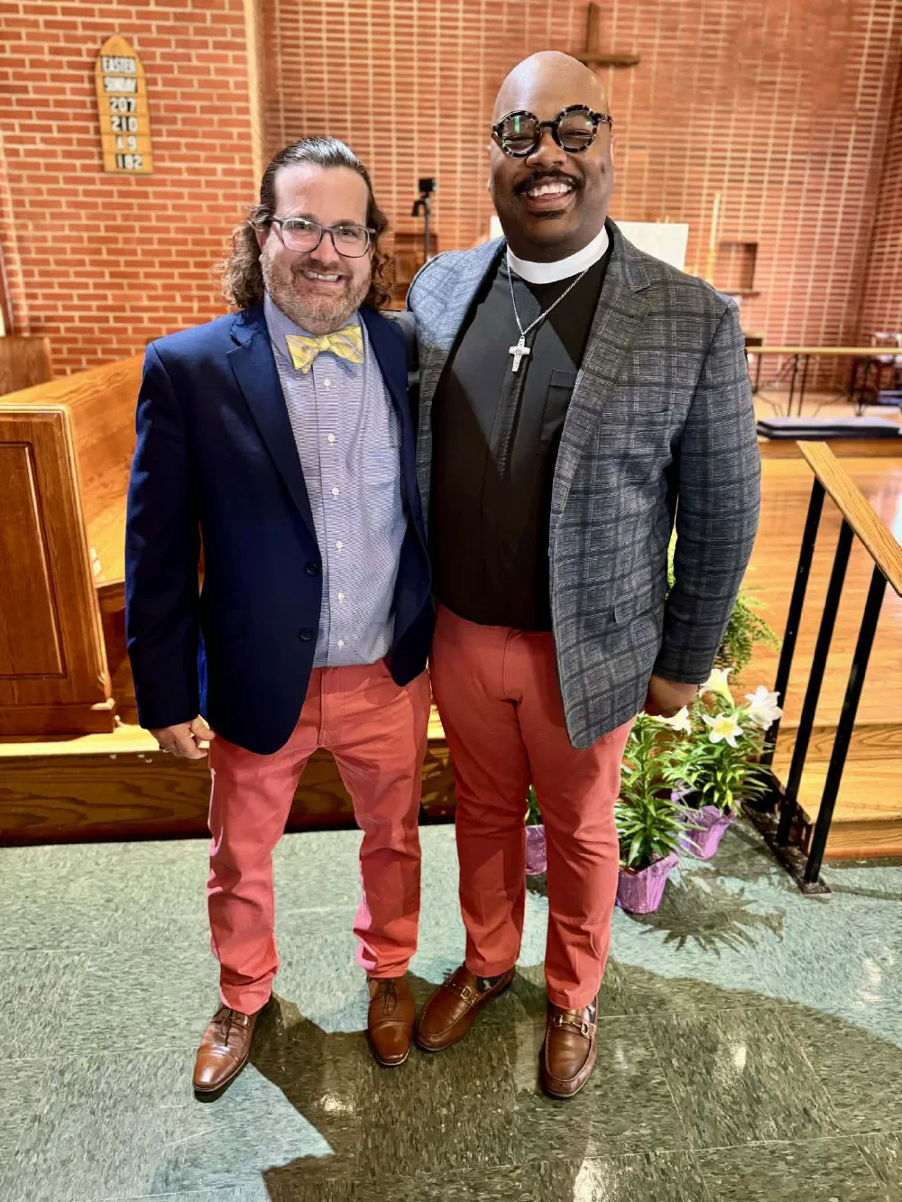 Two friends in matching coral pants, one in a bow tie and one in clericals, smile by the Easter lilies at the chancel steps
