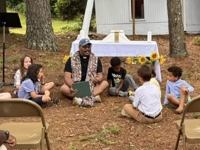 An outdoor altar dressed in white and flanked by sunflowers, with Father Quincy seated on the ground teaching the children