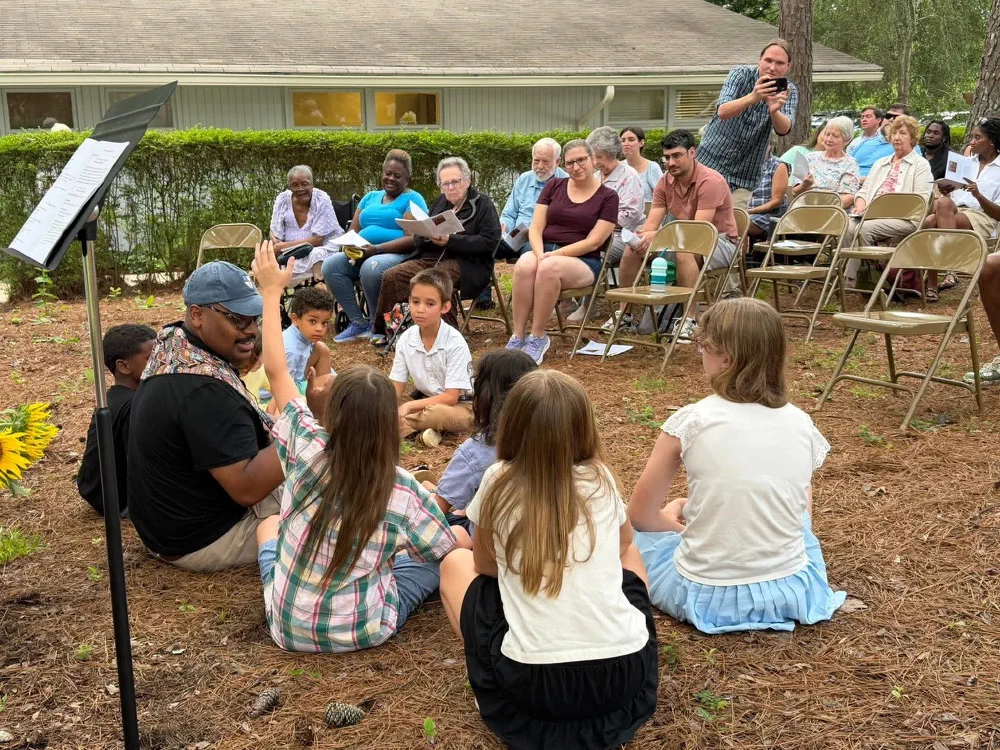 Father Quincy sits on the pine straw with children gathered in a circle while the congregation watches from folding chairs