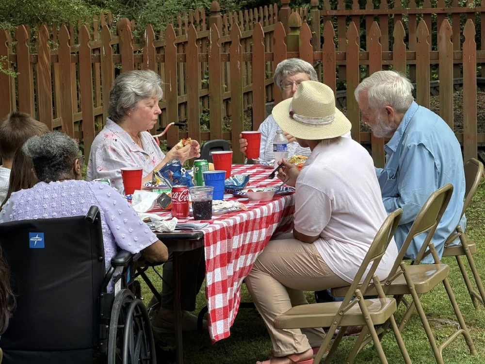 Older parishioners share lunch at a red checked table by the picket fence, one of them seated in a wheelchair