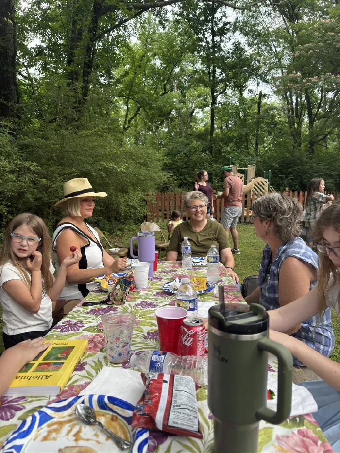Children and adults around a flowered picnic table crowded with cups, snacks, and a yellow Alleluia songbook