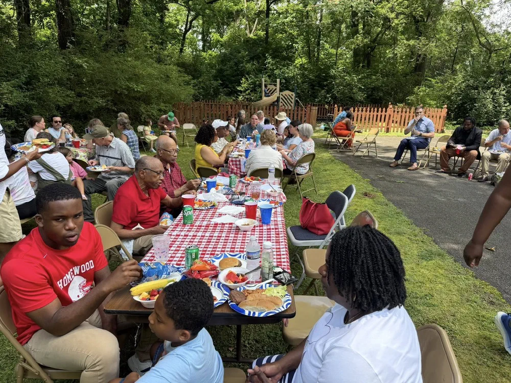Parishioners share lunch at long picnic tables with red checked cloths at the edge of the woods