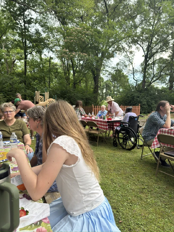 Families eating at picnic tables scattered across the grass beneath tall trees, with a playground behind
