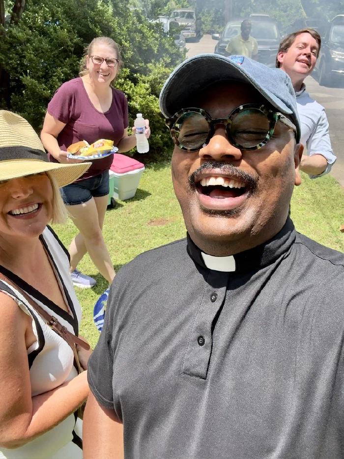 Father Quincy laughs into the camera in a sunny selfie with three smiling parishioners behind him