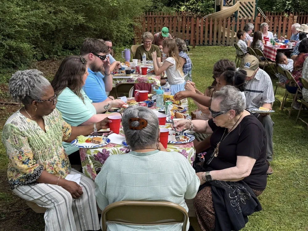 A full picnic table of parishioners of all ages passing plates under the trees