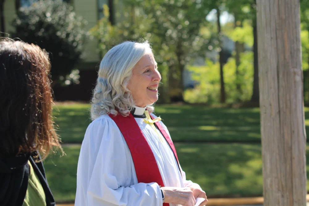 A priest in a white surplice and red stole smiles in the sunlight during the outdoor Palm Sunday liturgy